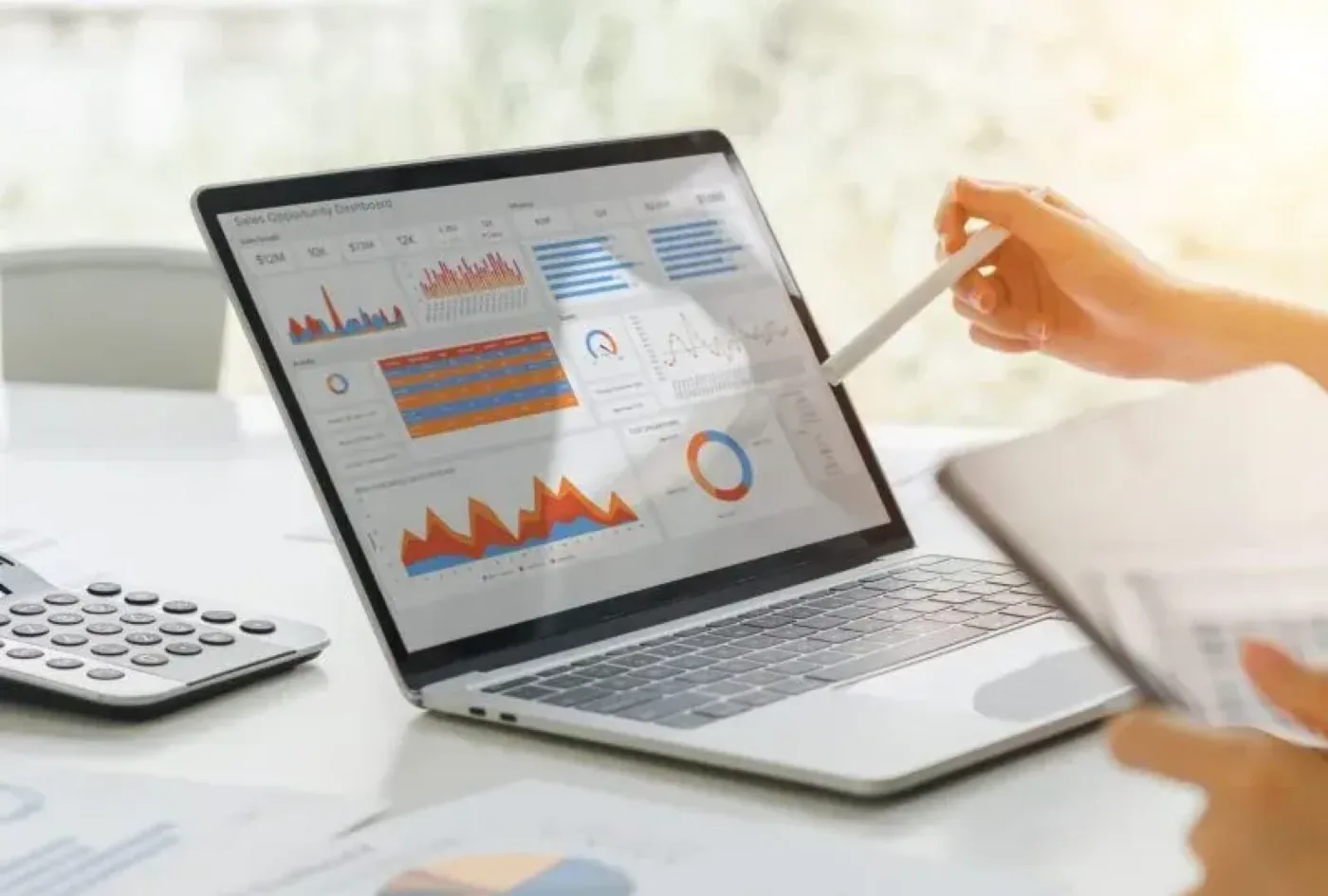 Notebook aberto em um dashboard de vendas com gráficos coloridos, enquanto uma mão aponta para a tela com uma caneta