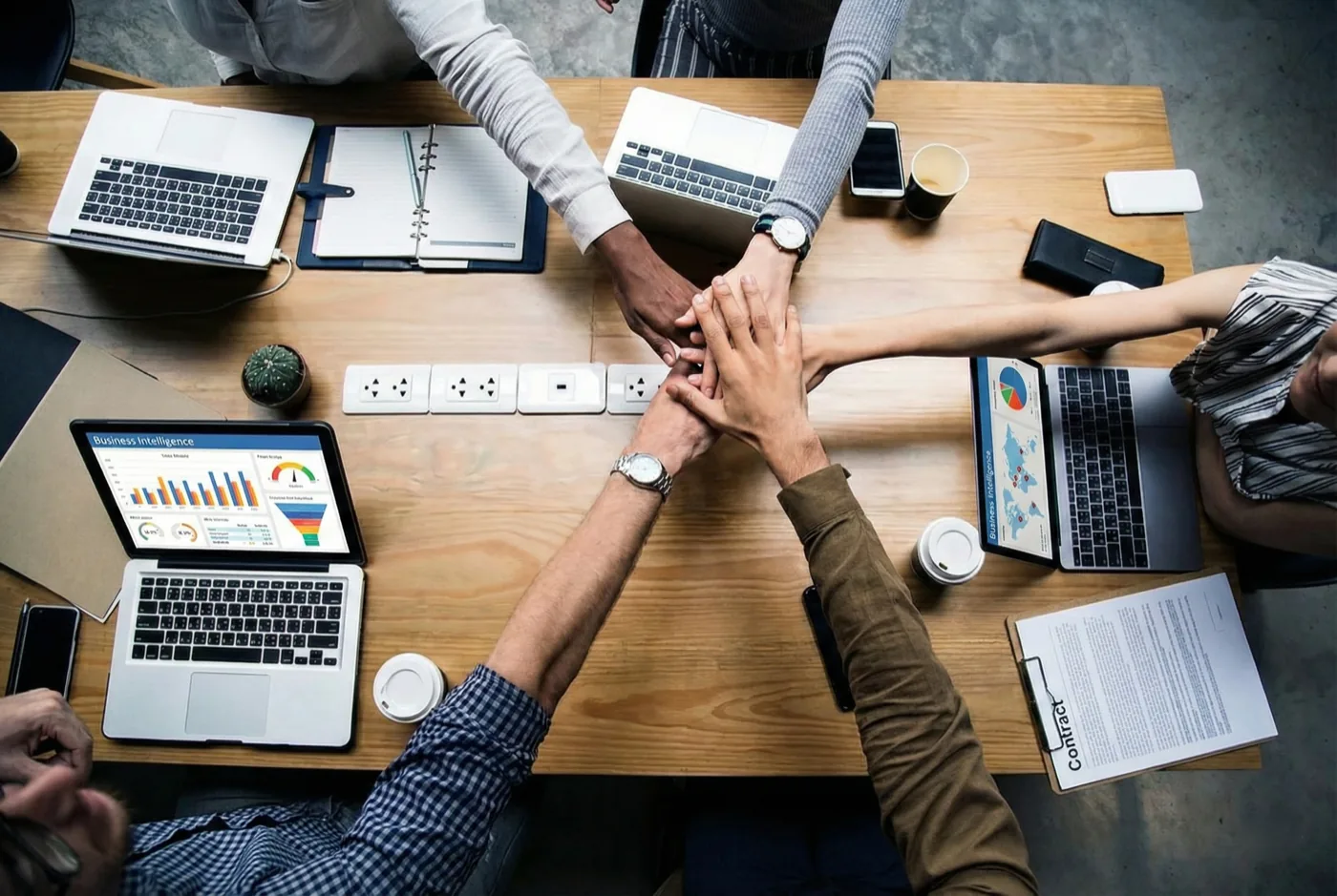 Equipe unindo as mãos sobre a mesa de reunião, cercada por notebooks com dashboards de Business Intelligence